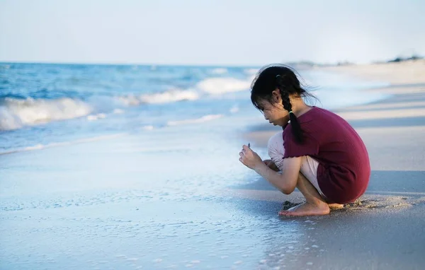 一个可爱的年轻亚洲女孩 坐在白色沙滩上 温水清澈 和家人度假时捡海贝 图库照片