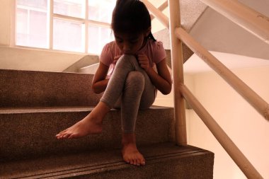Çocuk istismarı kavramı. Mutsuz ve yalnız bir genç kız, arka planda camdan içeri ışık saçan bir binanın içinde merdiven kenarında oturuyor..
