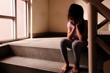 Çocuk istismarı kavramı. Mutsuz ve yalnız bir genç kız, arka planda camdan içeri ışık saçan bir binanın içinde merdiven kenarında oturuyor..