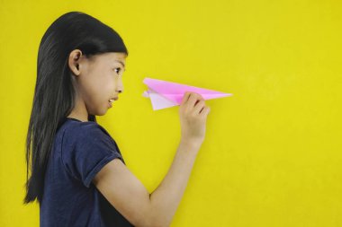 Genç ve sevimli bir Asyalı kız pembe kağıt uçağıyla oynuyor, tek eliyle tutuyor, nişan alıyor ve fırlatmaya hazırlanıyor. Parlak sarı arkaplan.