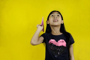 Genç ve sevimli bir Asyalı kız pembe kağıt uçağıyla oynuyor, tek eliyle tutuyor, nişan alıyor ve fırlatmaya hazırlanıyor. Parlak sarı arkaplan.