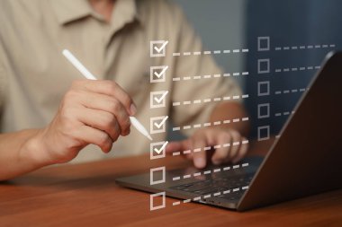 Dijital kontrol listesi ve görev yönetimi kavramı. Verimliliği, iş akışını, proje planlama ve verimlilik araçlarını sembolize eden sanal kontrol listesindeki öğeleri işaretlemek için dizüstü bilgisayarı olan bir kalem kullanan kişi.