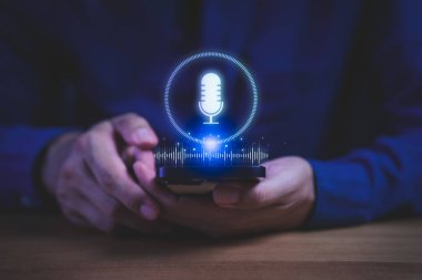 Parlak mikrofon ikonu ve ses dalgası hologramı ile akıllı telefon tutan kişi, ses tanımayı sembolize eden kişi, yapay zeka asistanı, dijital iletişim ve modern cihazlarda akıllı teknoloji yeniliği.