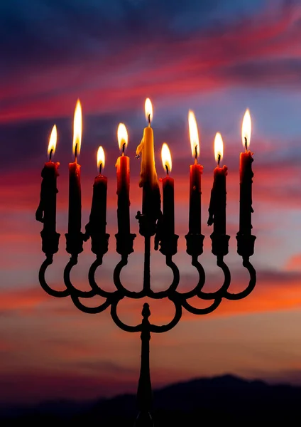 Glitter lights of candles on menorah are traditional symbols for Jewish Hanukkah Holiday of Light. Selective focus on candles. Background with blurred dramatic morning sky and mountains