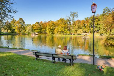 Bir çift mutlu son sınıf öğrencisi ve köpekleri eski sonbahar parkında bir gölet ya da nehir kenarında oturuyorlar.