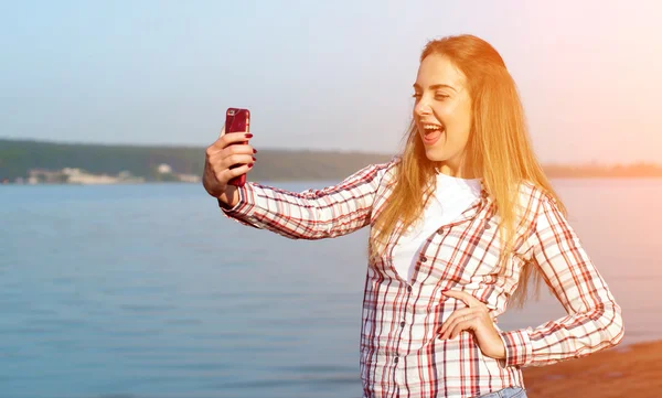 Beautiful emotional girl makes selfie near the lake at sunset