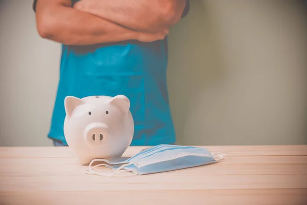 A white piggy bank with a blue medical mask on a wooden table. A man standing plan to save money during the coronavirus epidemic. Concept saving money on the future.