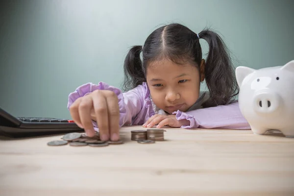 A cute Asian kid is counting money to drop the piggy bank with a black calculator placed on an old wooden table. Concept to save money for education future.