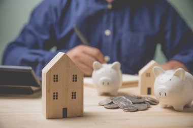 Paraları istiflenmiş ahşap ev modeline ve kumbarasına odaklan. İşadamı muhasebe kaydını hesap makinesi kullanarak gelecekte ev satın almak için hesaplama yapıyor. Konsept muhasebe, kredi satın almak için para biriktirme, iş.