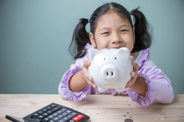 Concept to save money for education future. Cute Asian kid girl smile happiness holding white pig piggy bank with the calculator on wooden savings money for the cost of school supplies in the future.