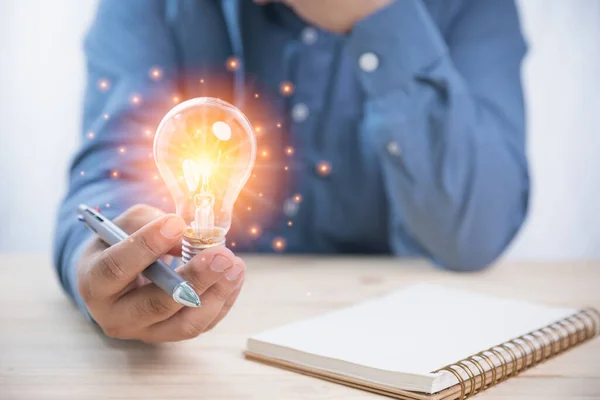 Concept business, innovation, ideas, and technology. A businessman holding a pen and a bright light bulb have a notebook on a wooden desk with copy space. Creativity innovation ideas for the future.