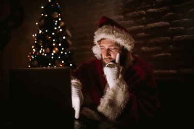 Noel Baba karanlık bir odada dizüstü bilgisayarın önünde otururken, Noel ağacının ışıkları arka planda görülebilir. Noel ve Yeni Yıl Kutlaması