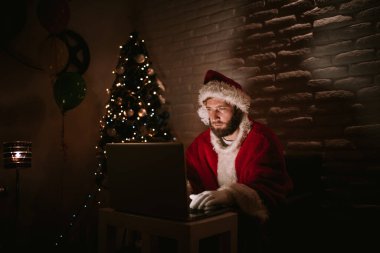 Noel Baba gibi giyinmiş yakışıklı beyaz bir adam, arka planında ışıklar olan bir Noel ağacı olan bir video görüşmesinde konuşuyor. Kış tatili