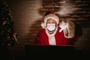 Noel Baba kostümü giymiş beyaz bir adamın portresi dizüstü bilgisayarın önünde oturuyor ve baş parmağını kaldırıyor. Yeni yıl kutlaması karantinada salgın COVID 'de - 19 koronavirüs