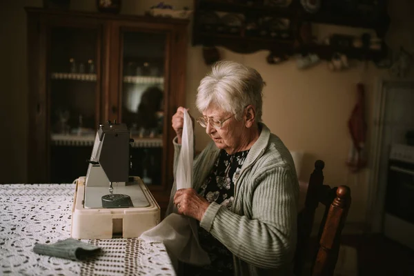 一位退休的高加索老妇人的侧视图 她手里拿着一块白布 手里拿着一副眼镜 在客厅里的缝纫机上缝纫 — 图库照片