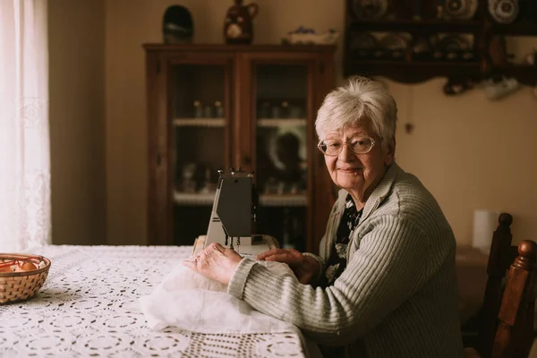一个带着退休眼镜的高加索祖母的画像 她在桌上缝纫机上缝上窗帘 退休老妇人的业余爱好 — 图库照片