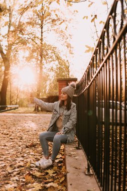 Kotlu örgü şapkalı genç bir kız parkta çitin üzerinde oturur ve telefonla selfie çeker. Parkta renkli bir sonbahar