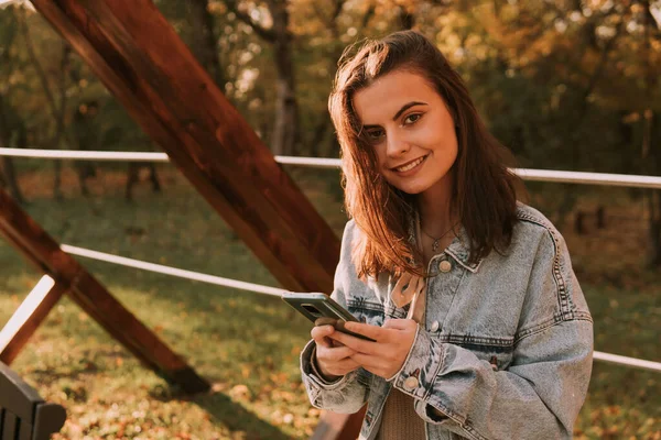 청바지를 소녀가 귀엽게 웃으며 벤치에 전화기를 사용하고 공원에서의 — 스톡 사진