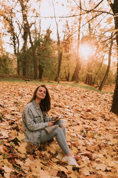 청바지를 아름다운 도시의 소녀가 공원의 나뭇잎에 전화를 가을에 공원을 — 스톡 사진