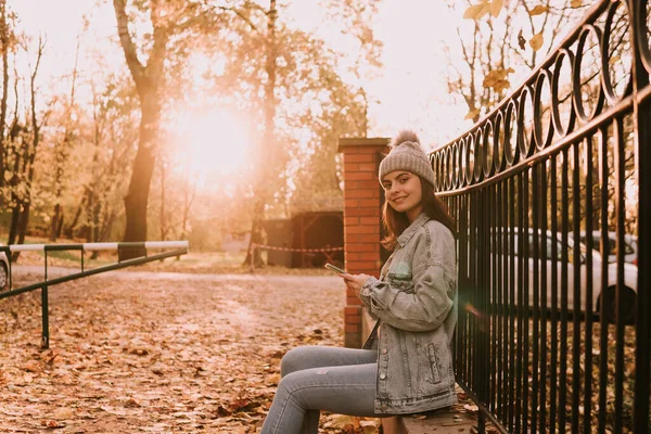 청바지를 울타리에 도시의 소녀가 휴대폰으로 메시지를 입력하고 카메라를 공원의 — 스톡 사진