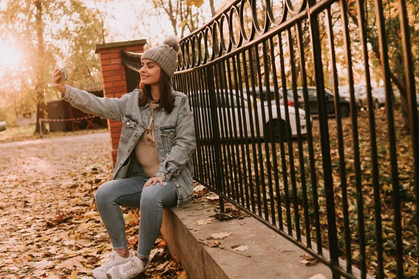 청바지를 모자를 소녀가 울타리에 전화로 셀카를 공원의 — 스톡 사진