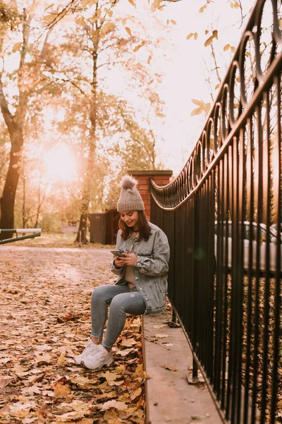 청바지를 현대의 아름다운 소녀가 울타리에 스마트폰을 가을에 공원을 거닐고 — 스톡 사진