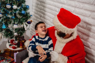 Noel Baba kostümü giymiş genç sakallı bir adam kollarında tuttuğu güzel bir çocuğa odada süslü mavi bir Noel ağacının yanında gülüyor. Mutlu yıllar ve mutlu yıllar.