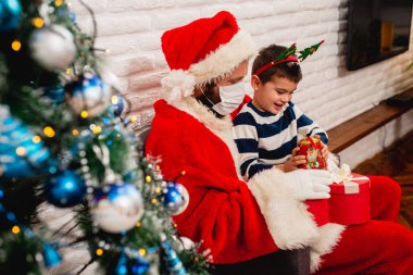 5 yaşında güzel bir çocuk Noel Baba 'nın kucağında oturmuş, maskeli ve odada hediyesini seyrediyor. COVID salgını sırasında kış tatili -19 koronavirüs