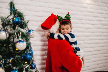 Noel Baba kostümü giymiş bir adamın kollarında kafasına aksesuar takmış sevimli bir çocuk evde süslü Noel ağaçları giyiyor. Yılbaşı ve Noel evde