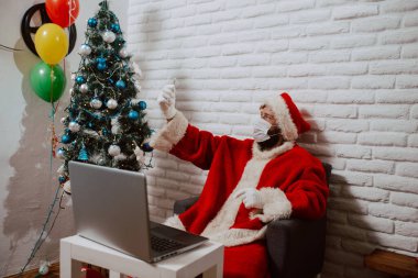Noel Baba kostümü giymiş, tıbbi maske takan genç bir adam evde dizüstü bilgisayarın önünde oturur, selfie çeker ve bir video görüşmesine onay verir. Noel Baba COVID sırasında çevrimiçi - 19 