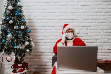 Noel Baba kostümü giymiş sakallı genç bir adam evde dizüstü bilgisayarın önünde oturuyor ve telefonla konuşuyor. COVID sırasındaki yılbaşı ve xmas - 19 koronavirüs
