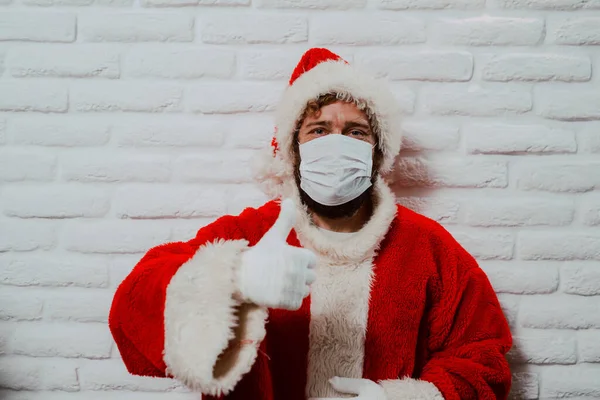 A portrait of a young man with a face mask dressed in a Santa Claus suit standing in front of a white wall and giving a thumbs up. Holidays at home during COVID - 19 coronaviruses