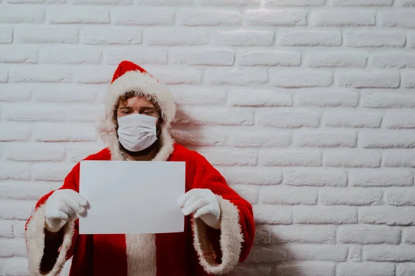 A portrait of a bearded young man with a face mask dressed in a Santa Claus suit standing in front of a  wall in the house and holding an empty white banner. Holidays during COVID - 19 coronavirus