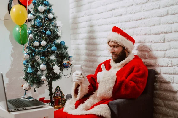 산타클로스 남자가 태블릿을 의자에 위에는 크리스마스 트리가 받으세요 — 스톡 사진