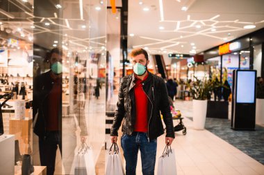 Siyah deri ceketli, alışveriş merkezindeki bir butik vitrinin önünde elinde maske ve çanta olan beyaz bir adam COVID virüsü salgını sırasında - 19 Coronavirus