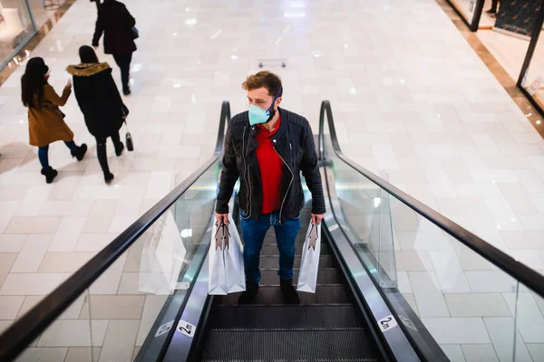 An urban young man with bags in his hand and a medical face mask on the escalator in a shopping mall. Shopping during the COVID pandemic - 19 coronavirus