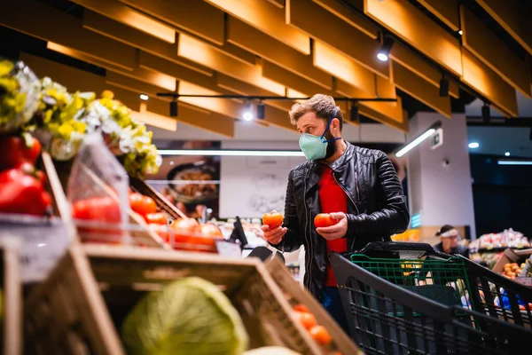 A handsome young man with a face mask buys a tomato at the supermarket. Procurement of food during the coronavirus crisis COVID - 19 pandemic