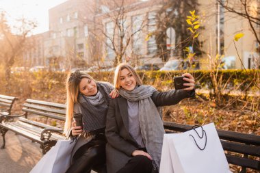 Uzun dalgalı saçlı, güzel bir sarışın kızın portresi bankta oturmuş bir şeyleri tartışıyor, önlerinde alışveriş torbaları ve selfie çekiyor.