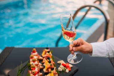 Kadın model havuz kenarında çilekli bir bardak şarap tutuyor ve arka planda da şamdanlar var.