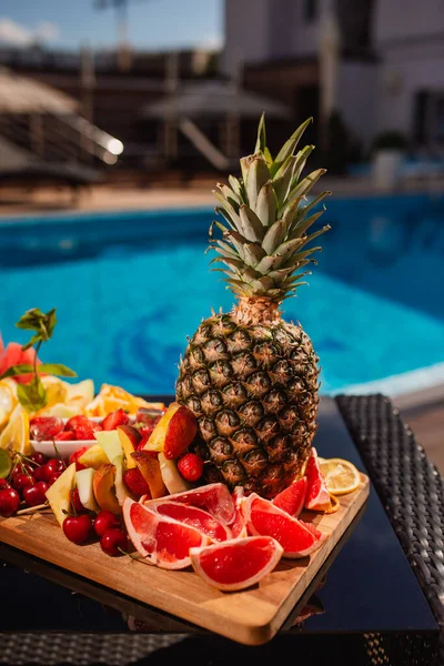 Tropikal meyveler suyun yakınındaki bir tabakta yatıyor. Ejderha meyvesi, papaya, çarkıfelek meyvesi, mango, mavi yüzme havuzunun arka planında ananas. Yakın çekim.