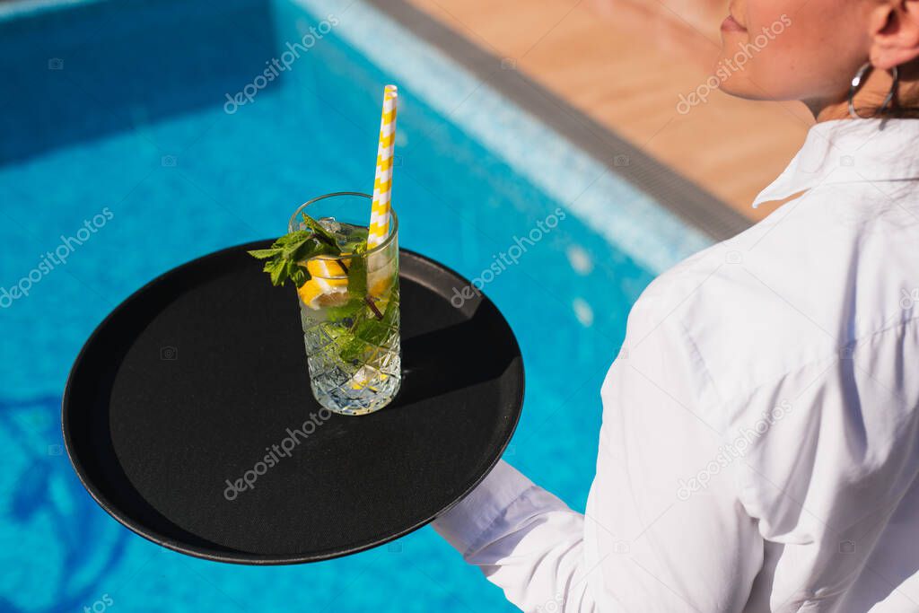 A waitress in a white shirt serves a mojito cocktail to guests by the pool. Outdoor summer party