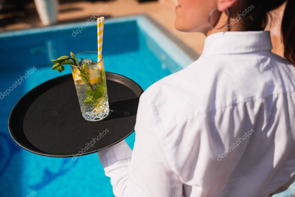 A female waitress serves a mojito cocktail on a tray by the pool. Crazy summer party