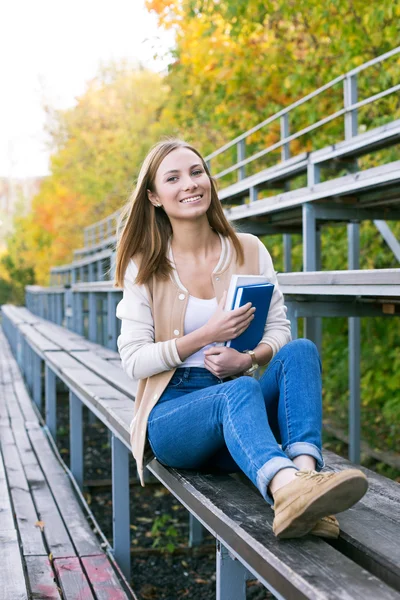 Estudante sentado no tribuno do esporte com livro e sorrindo — Fotografia de Stock