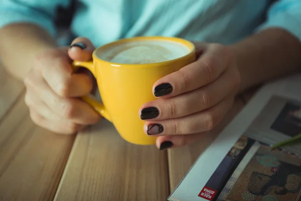 Yellow cup in the hands of women. Gel nail manicures. Phone and the magazine on the table