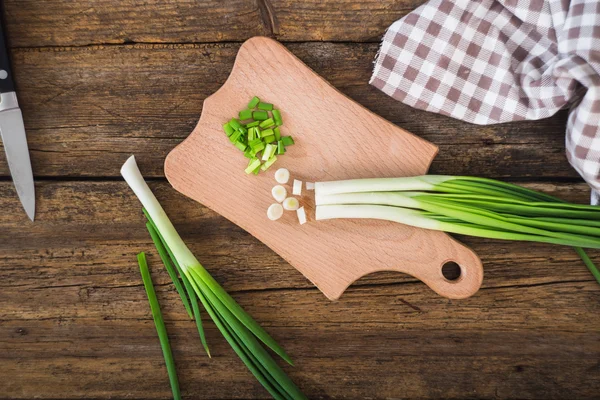 Grüne Zwiebeln auf einem Schneidebrett. Messer und Serviette auf einem Holztisch — Stockfoto