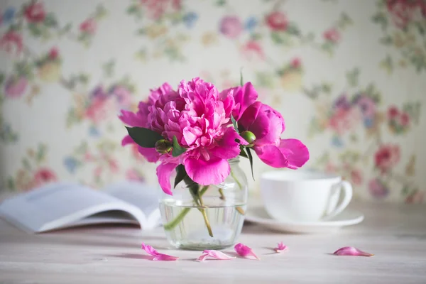 Beyaz ahşap bir masa üzerinde bir cam kavanoza peonies taze buket. kitap ve bir fincan çay