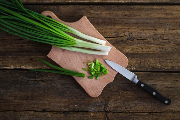 Cebolas verdes em uma tábua de cortar. Faca e guardanapo em uma mesa de madeira — Fotografia de Stock