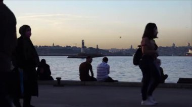 Kadıköy / İstanbul: Kalabalık sahil kenarı. İnsanlar oturur, deniz kenarında yürür. Feribotlar arkadan geçiyor