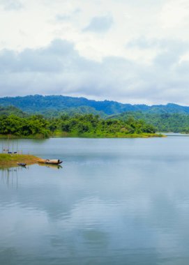 Bulutlu bulutlu yeşil dağlarla çevrili güzel bir göl manzarası. Kıyıda dinlenen bir tekne. Rangamati, Bangladeş 'te.