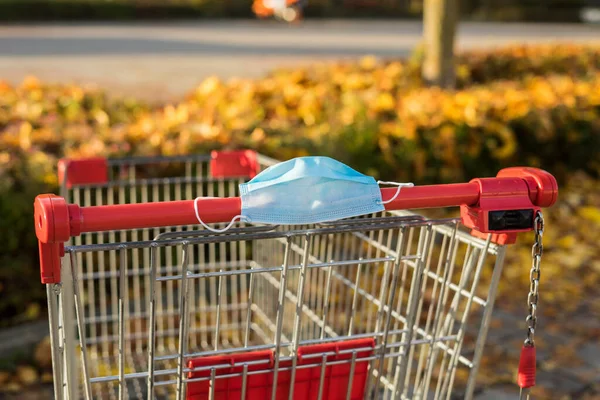 Grocery shopping with face mask during the coronavirus COVID-19 pandemic. Empty grocery shopping cart outdoor. Autumnal blurred background. Go shopping with a medical face mask.